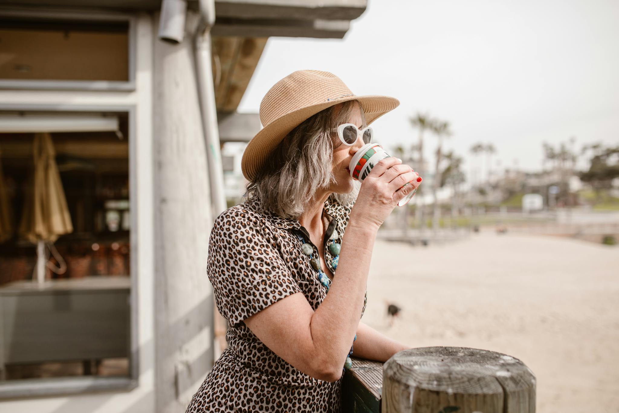 Stylish woman in leopard print and hat enjoying coffee outside a cafe by the beach.